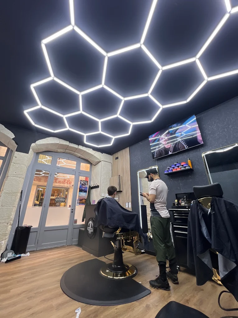 Barbier en plein travail chez L'Art du Barber à Forcalquier — fauteuil noir et doré, plafond LED hexagonal